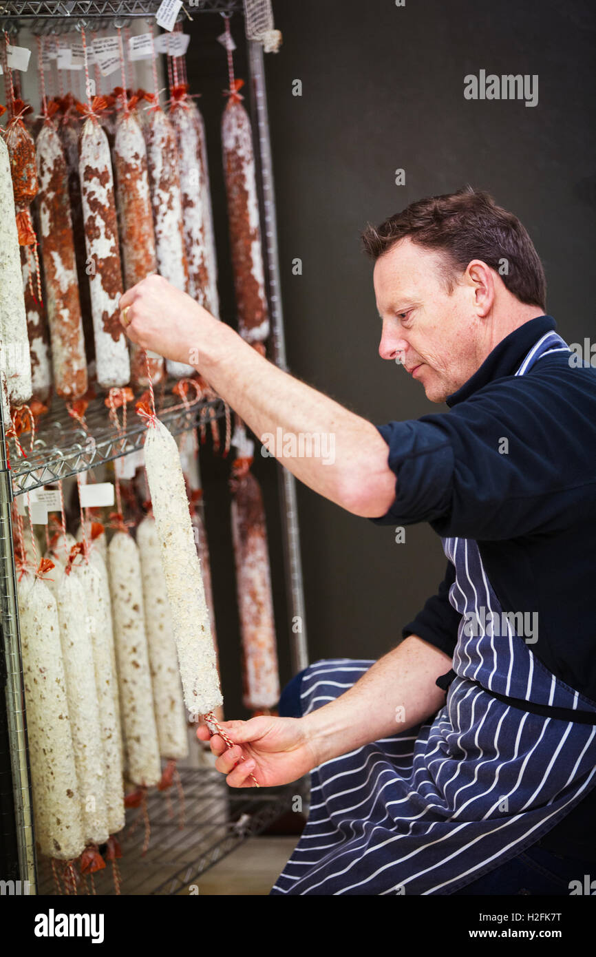 Metzger trägt eine gestreifte blaue Schürze Salamis auf einem Wäscheständer hängen. Stockfoto