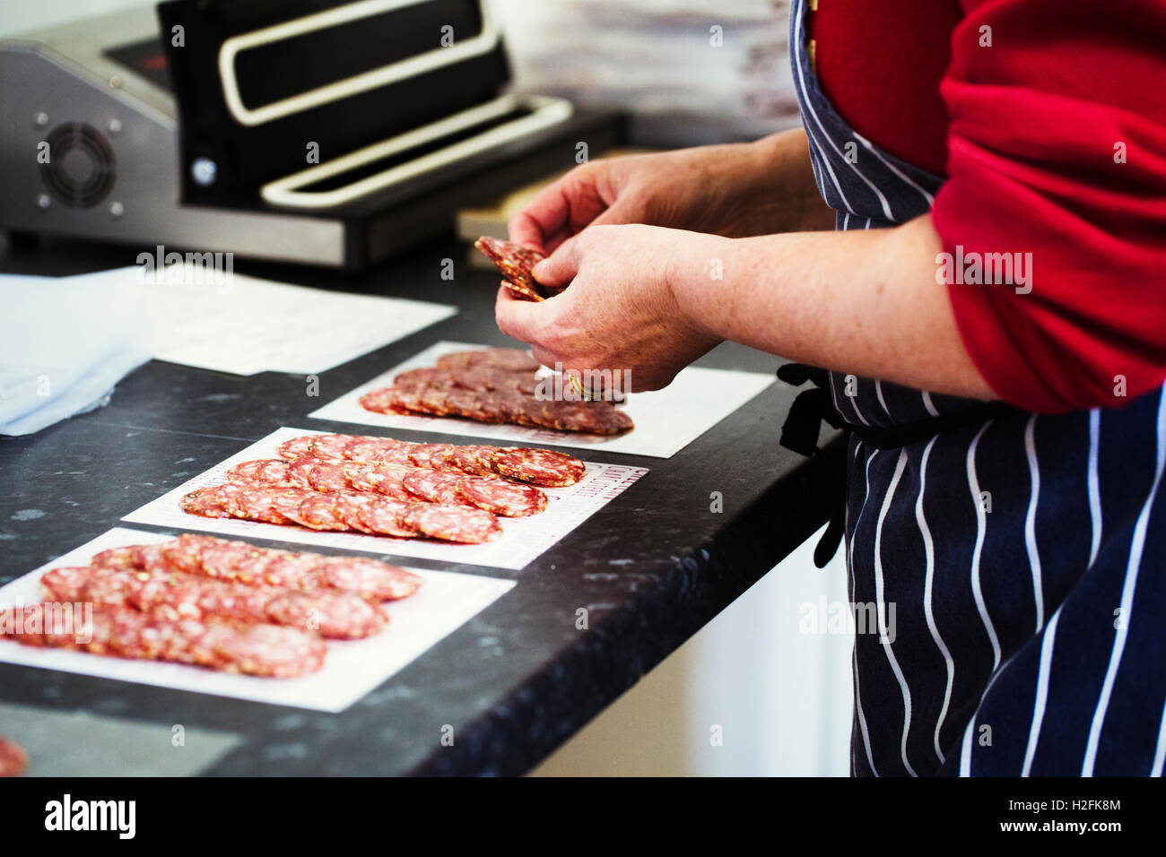 Nahaufnahme eines Metzgers trägt eine gestreifte blaue Schürze Verpackung Scheiben Salami. Stockfoto