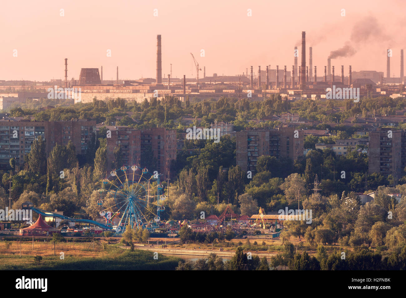 Vergnügungspark mit Riesenrad und Gebäude auf dem Hintergrund der Metallurgie Pflanze mit Smog bei Sonnenuntergang. Ökologie-Probleme, atmo Stockfoto