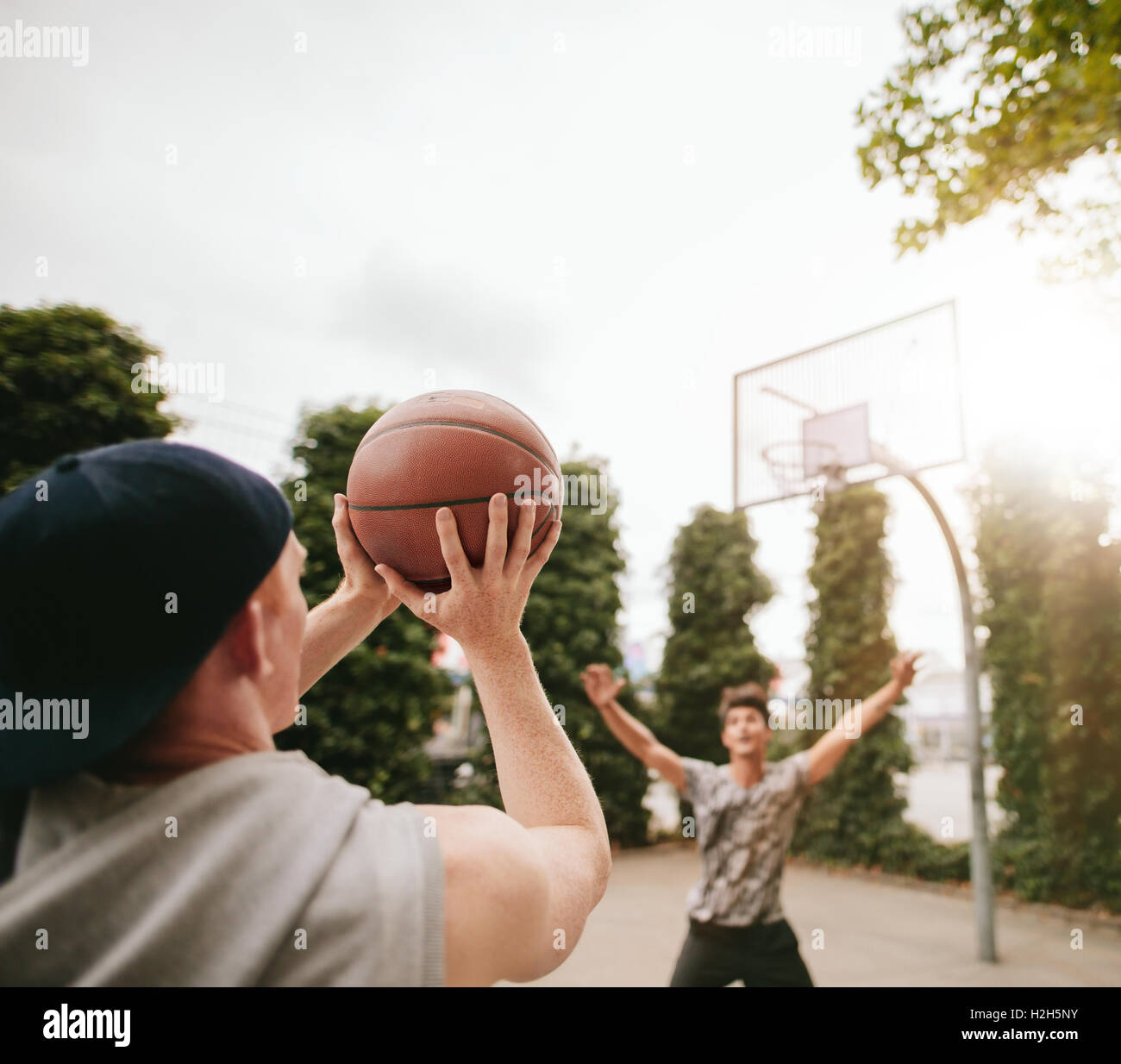 Junger Mann schießt den Ball in den Korb mit Freund im Hintergrund blockiert. Freunde spielen Basketball gegeneinander an heraus Stockfoto