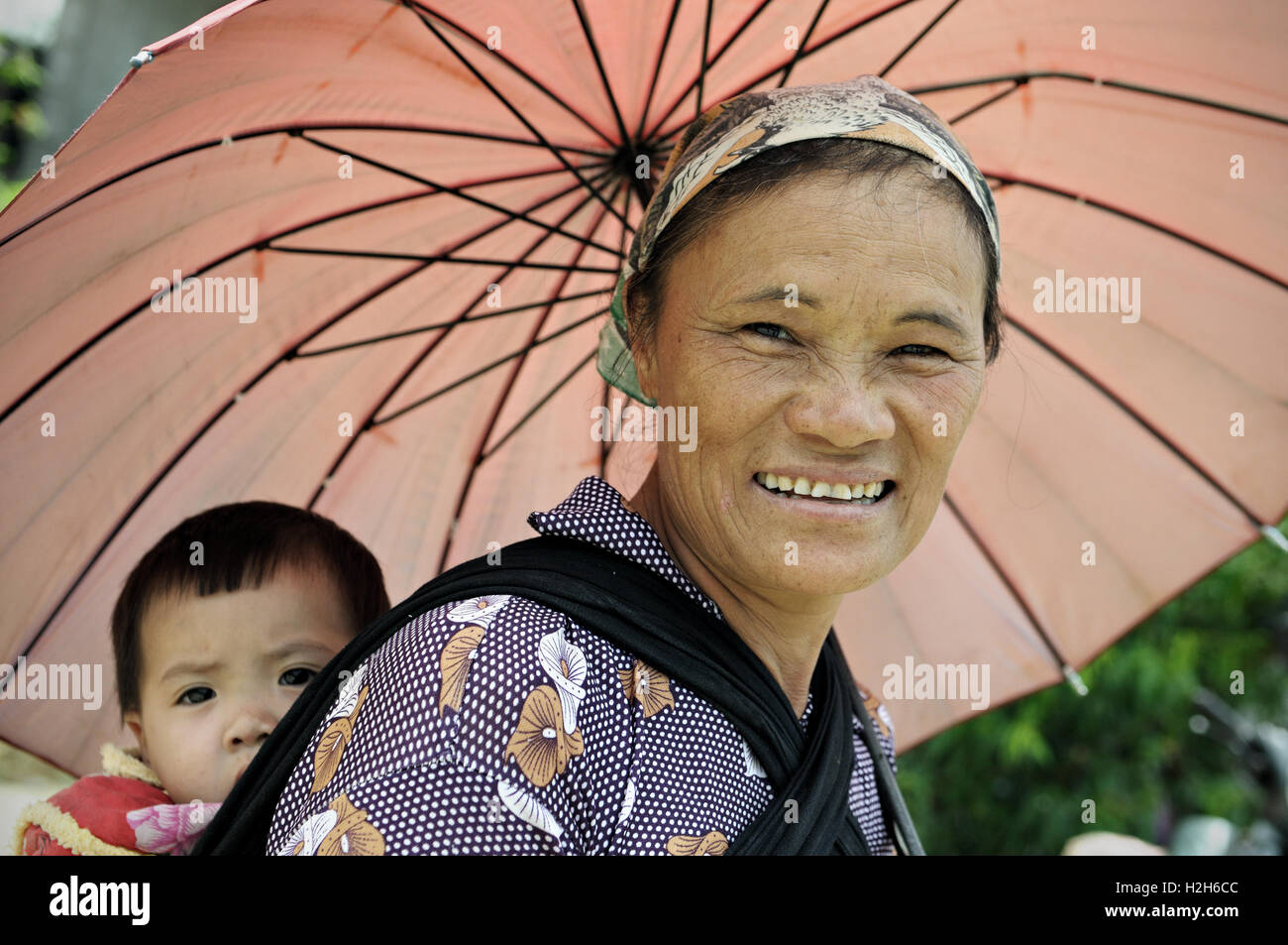 Frau mit Regenschirm tragen ein Baby auf dem Rücken auf dem Quyet Tien-Markt in Ha Giang Provinz, Nord-Vietnam Stockfoto