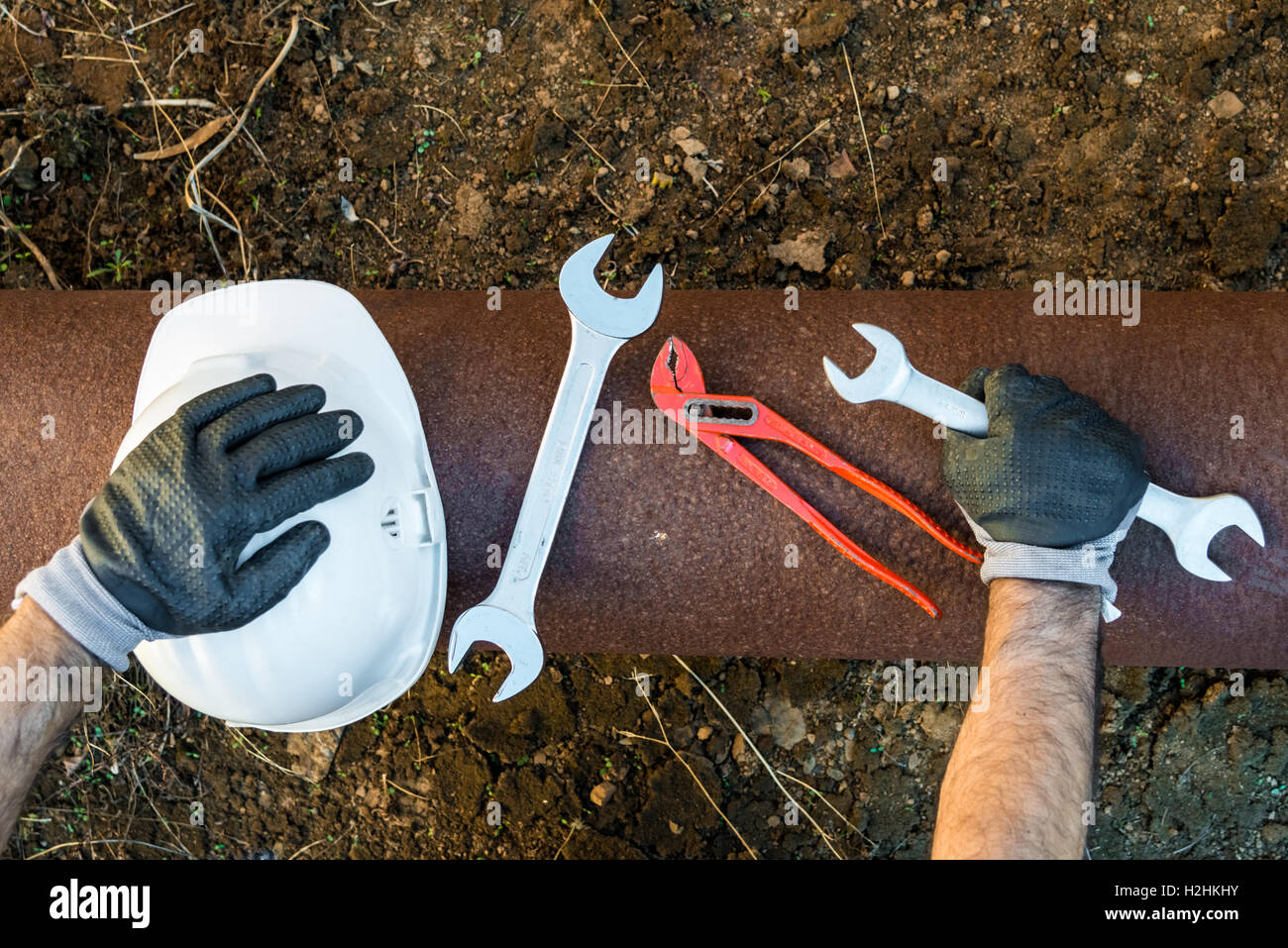 Hände mit Arbeitshandschuhen hält einen Schutzhelm, einen Schraubenschlüssel und einen roten Schlüssel Rohr klemmt Stockfoto