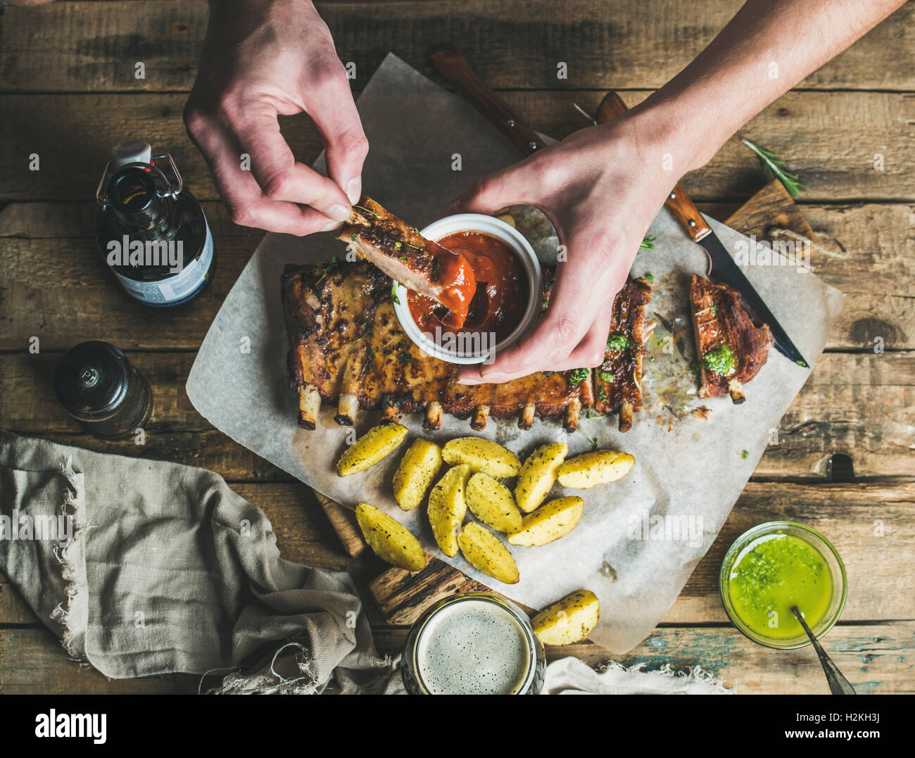 Menschen essen gebratene Schweinerippchen mit Kartoffeln, Knoblauch, Rosmarin und grüner Kräutersauce auf rustikalen Holztisch. Mann ' s Hände Dipp Stockfoto