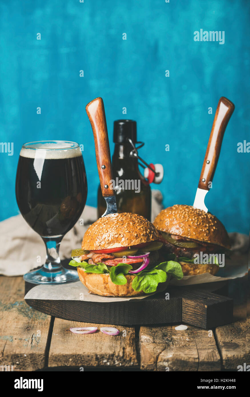 Zwei hausgemachte Rindfleisch-Burger mit knusprigem Speck, Zwiebeln, Gurken, Gemüse, Glas und Flasche dunkles Bier auf Holzbrett über Rost Stockfoto