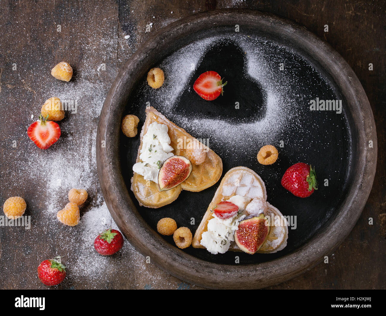 Wafer als Herzform mit gelbe Himbeeren, Erdbeeren, in Scheiben geschnittene Feigen, Ricotta-Käse und Zucker Pulver auf Ton Fach über dunkel Stockfoto