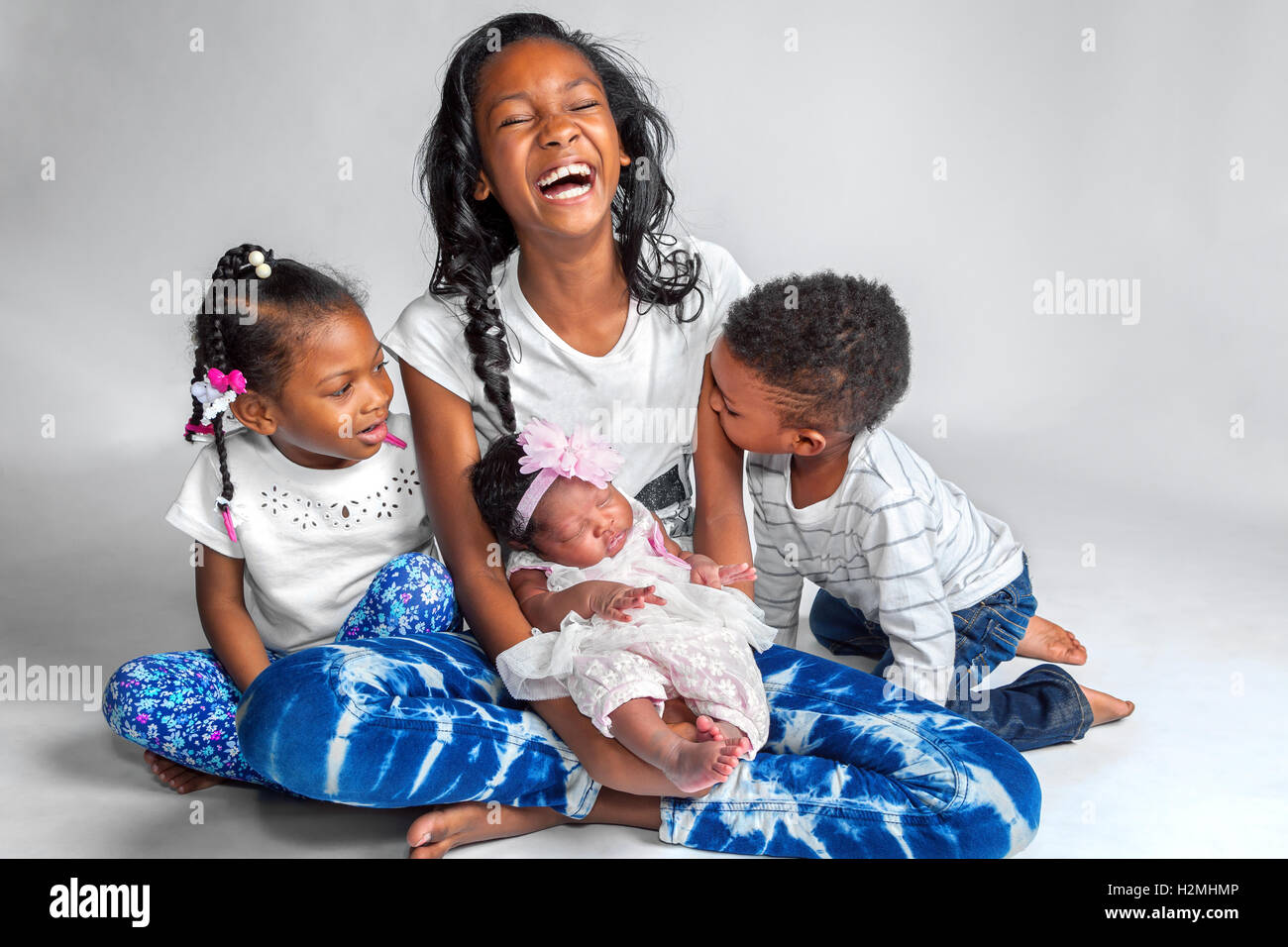 Vier afroamerikanische Geschwister sitzen auf einem weißen Hintergrund für ein Porträt.  Ein lachender Tween hält ihre Babyschwester während h Stockfoto