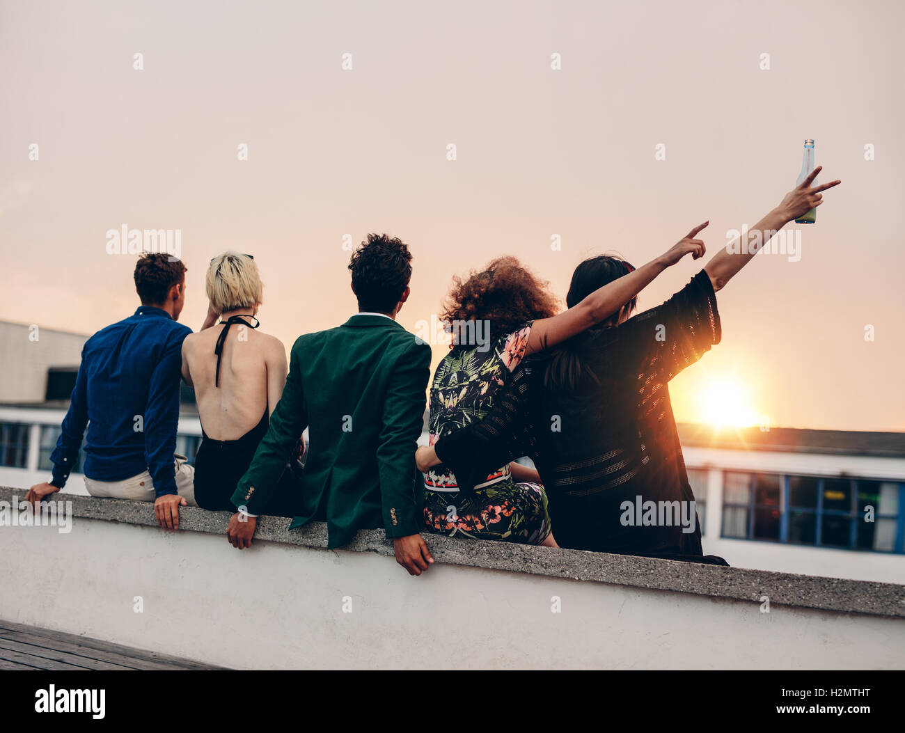 Gruppe von Freunden feiern auf Terrasse mit Getränken. Junge Männer und Frauen genießen Drinks auf der Dachterrasse bei Sonnenuntergang. Stockfoto