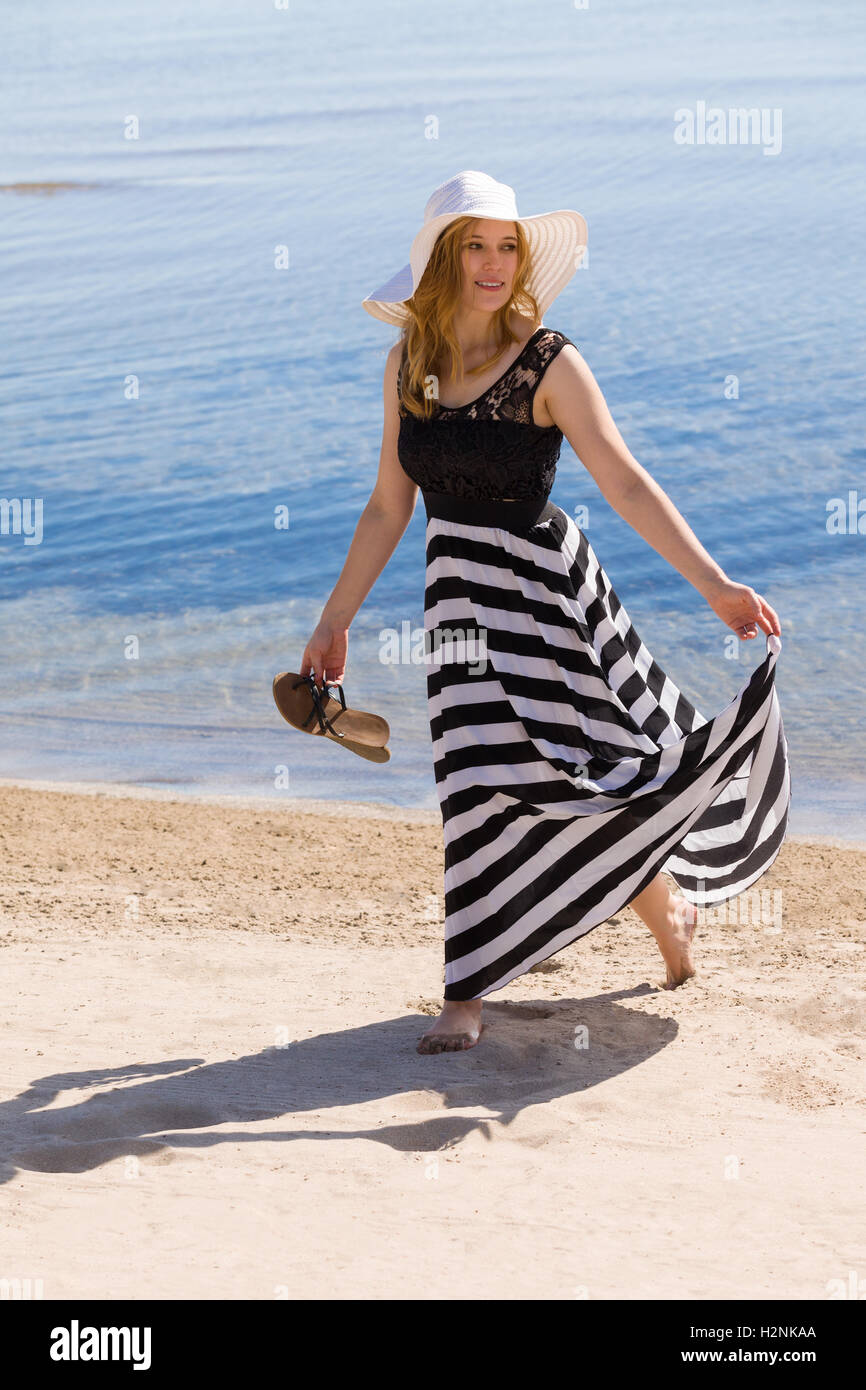 Schöne Frau in einer schwarzen und weißen Sonne-Kleid am Strand entlang spazieren. Stockfoto