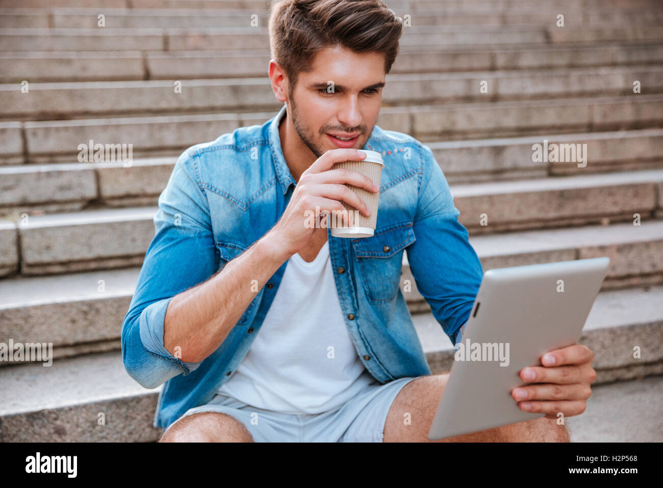 Zuversichtlich, dass junge Mann trinken nehmen Weg Kaffee und Blick auf pc tablet beim Sitzen auf der Treppe im freien Stockfoto