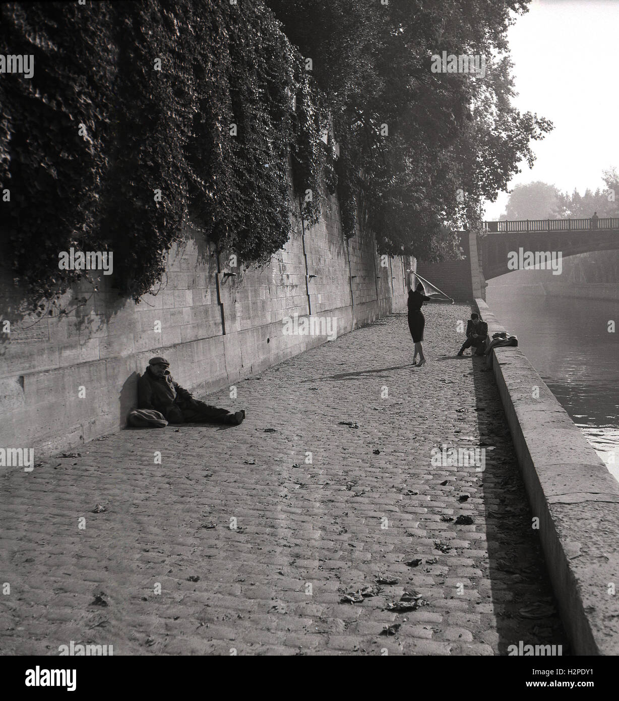 1950er Jahre, eine Fashion Shooting am Ufer der Seine, Paris, Frankreich, Fotografen, die Bilder von einer Frau in Heels und Kleid, mit einem vagrant sitzt auf dem Kopfsteinpflaster gegen eine Wand. Stockfoto