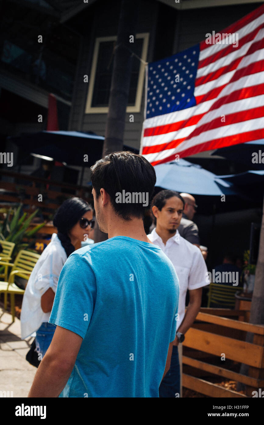 Junger Mann im blauen Hemd entlang einer Straße in La Jolla, Kalifornien mit Sonnenschirmen und eine amerikanische Flagge im Hintergrund. Stockfoto