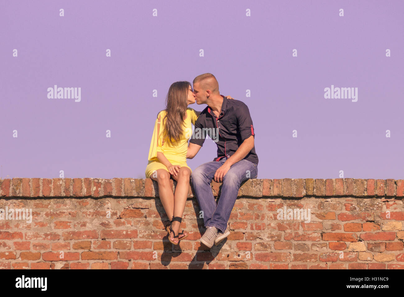 junge Erwachsene 20 s paar küssen Sitzung Ziegel Wand lila Himmel Stockfoto