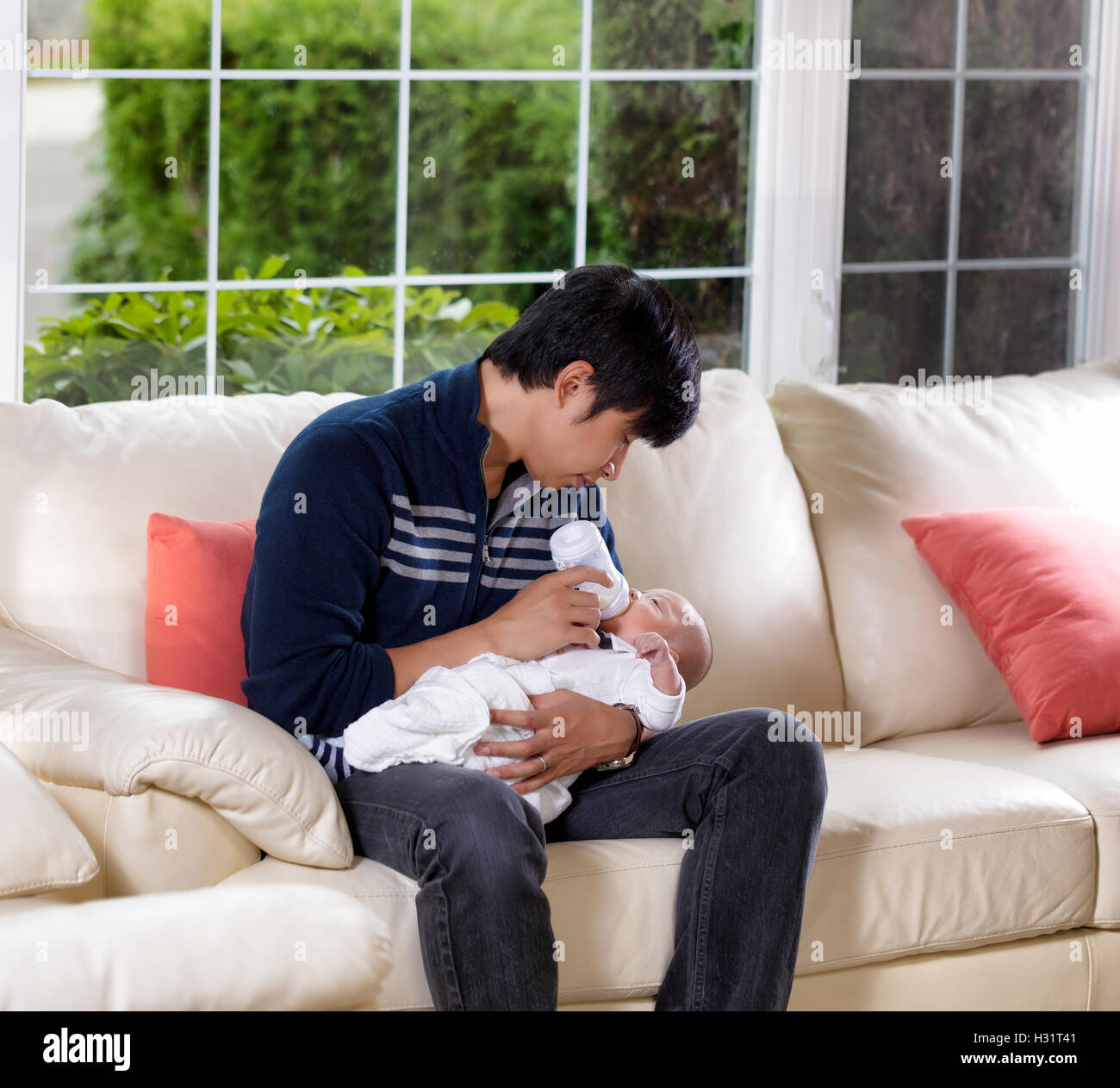 Vater Fütterung junges Baby Säugling junge Familie weißen Sofa mit großen Fenstern im Hintergrund Stockfoto