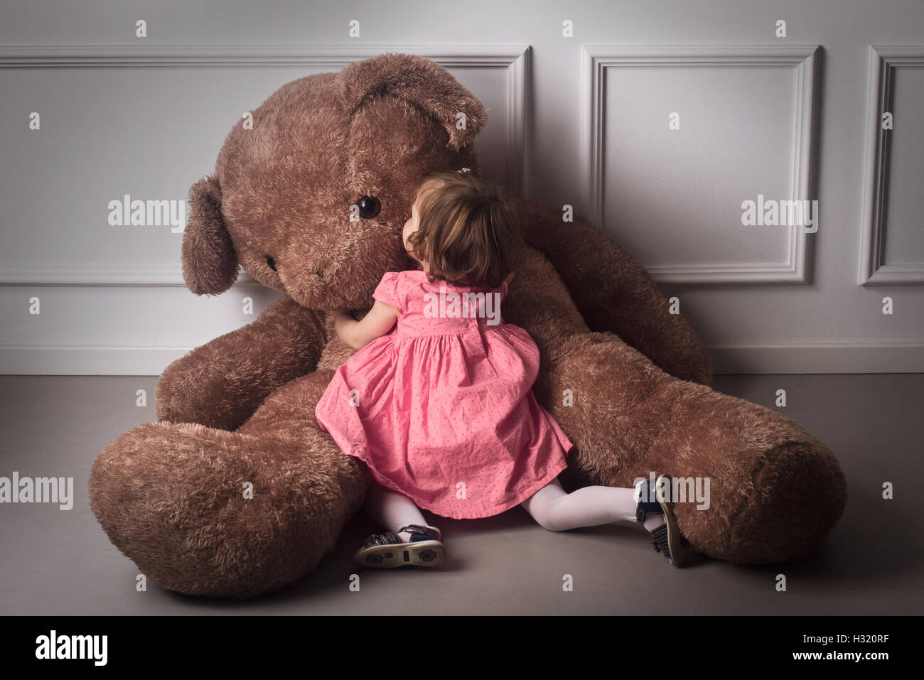 Horizontale Rückansicht von kleinen Mädchen in Ping Kleid umarmt einen sehr großen Teddybär auf dem Boden sitzend Stockfoto