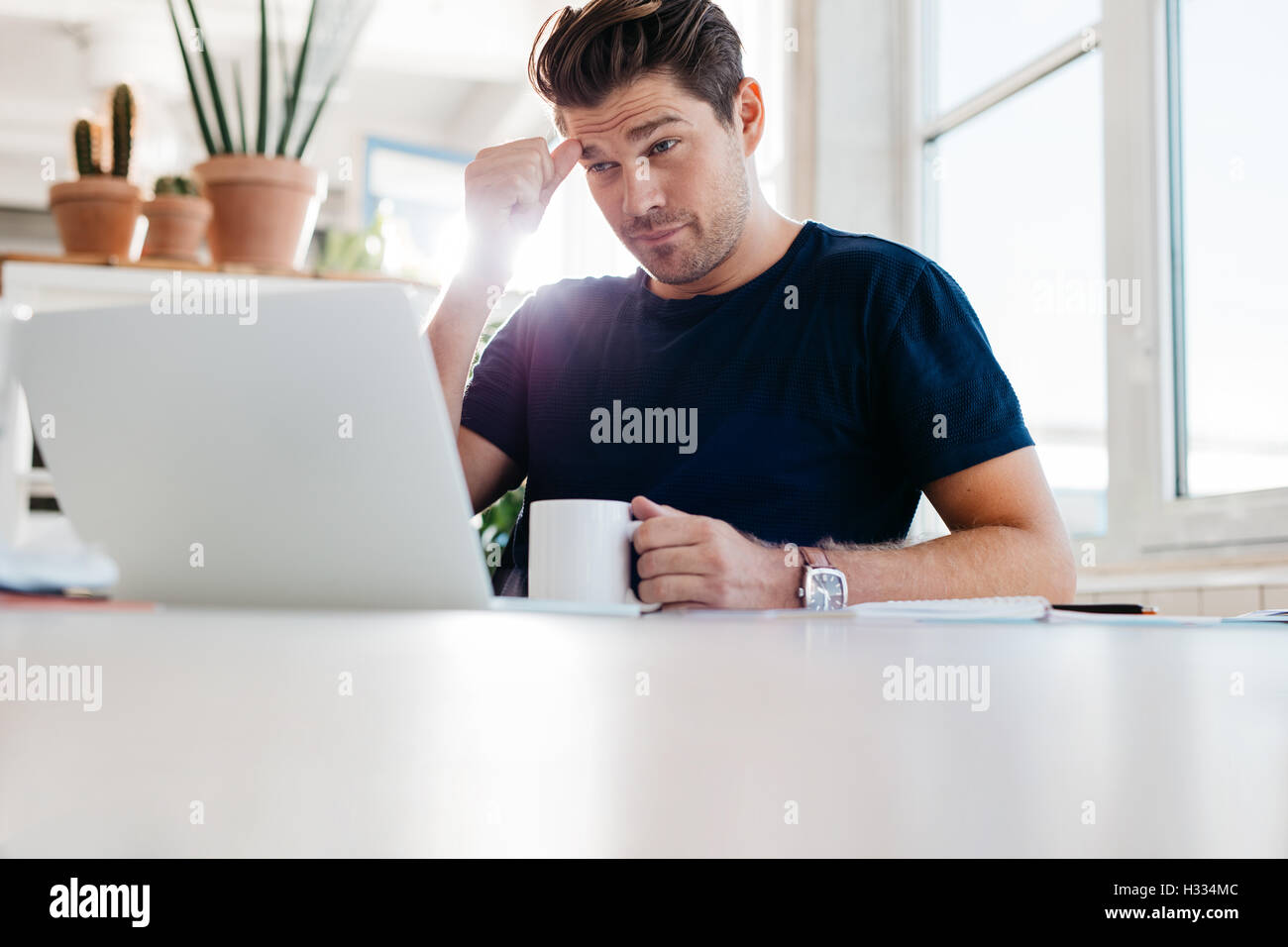 Nachdenklicher junger Mann Kaffeetasse halten und mit Blick auf Laptop sitzend an seinem Arbeitsplatz im Büro. Stockfoto