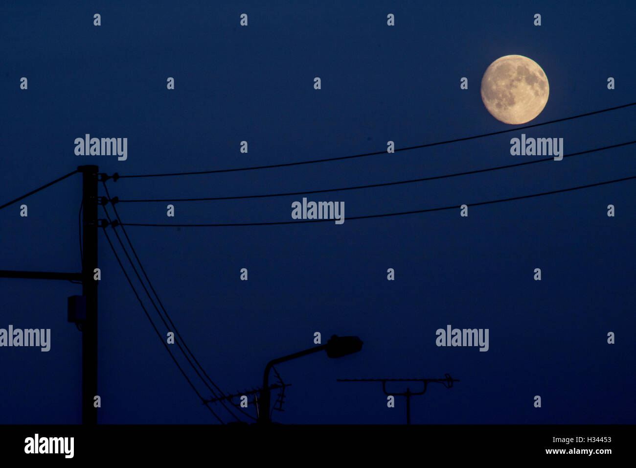 Vollmond im blauen Himmel über Telefonleitungen mit Silhouetten von Straßenlaternen Stockfoto