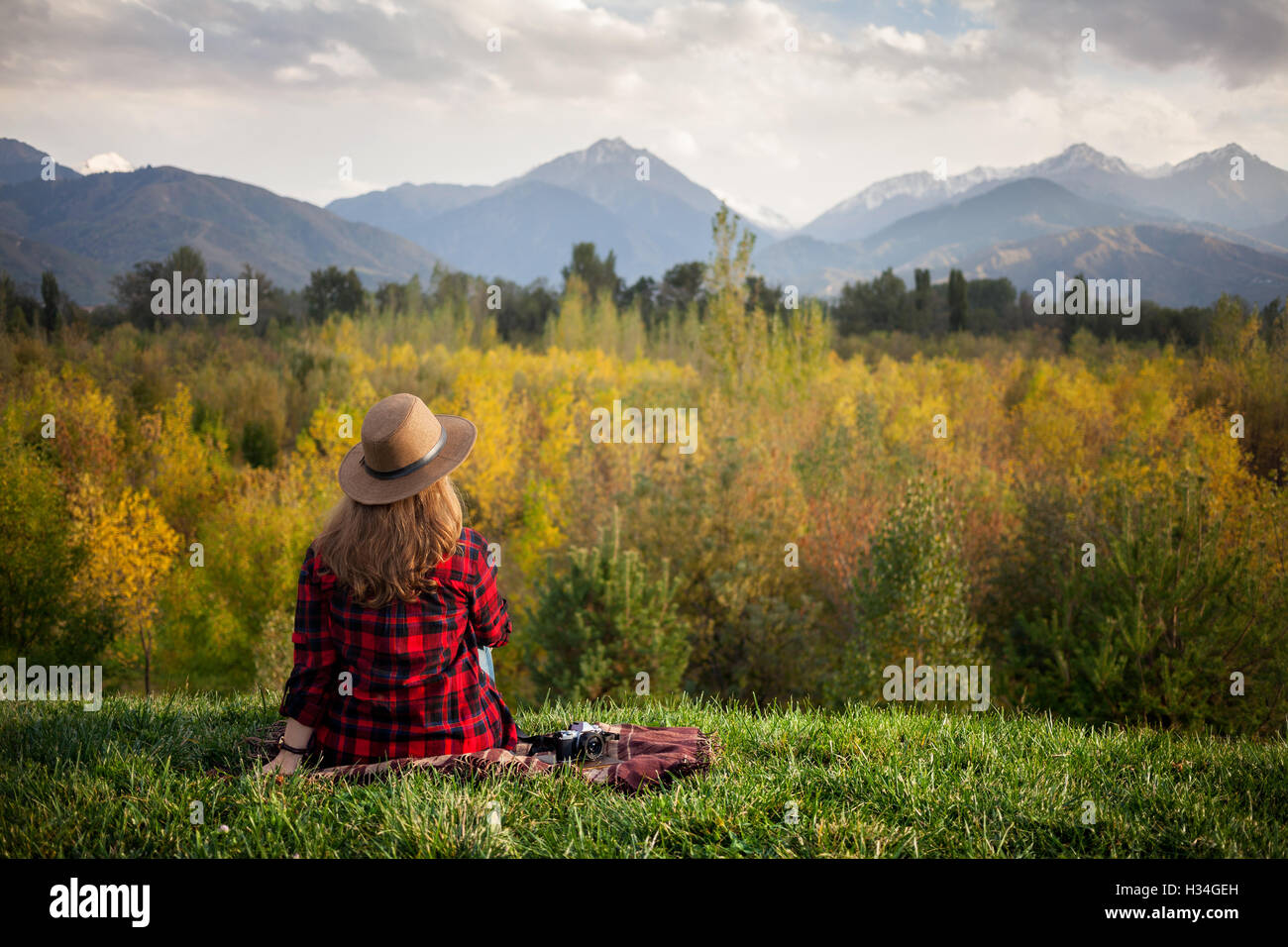 Frau in Rot kariertes Hemd und Hut sitzen auf dem Rasen mit Fotokamera im herbstlichen Wald und Berge-Hintergrund Stockfoto