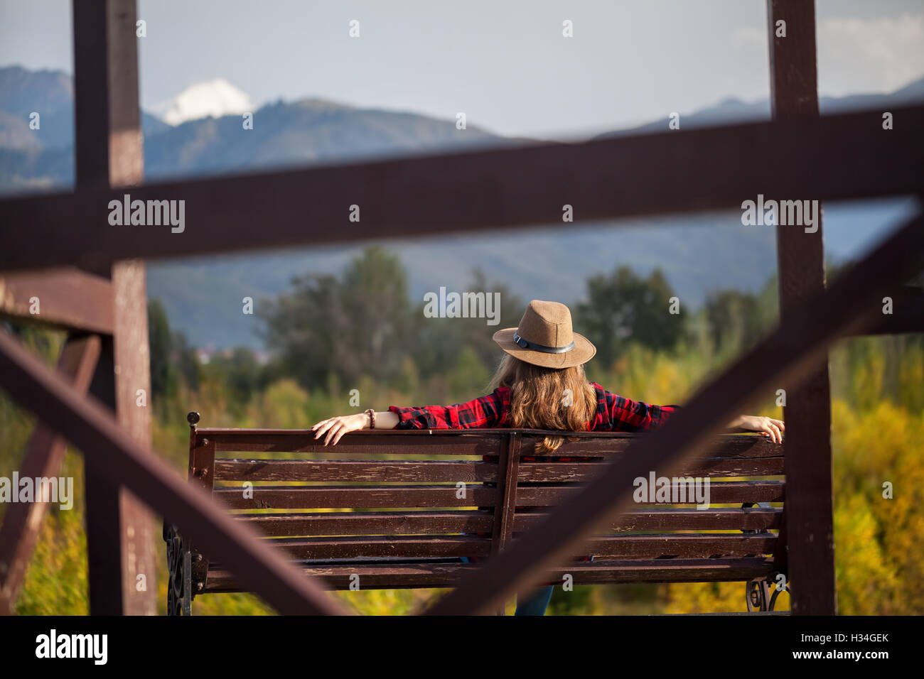 Frau in Rot kariertes Hemd und Hut sitzt auf der Holzbank im herbstlichen Wald und Berge-Hintergrund Stockfoto