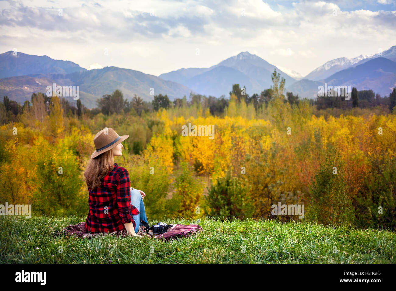 Frau in Rot kariertes Hemd und Hut sitzen auf dem Rasen mit Fotokamera im herbstlichen Wald und Berge-Hintergrund Stockfoto