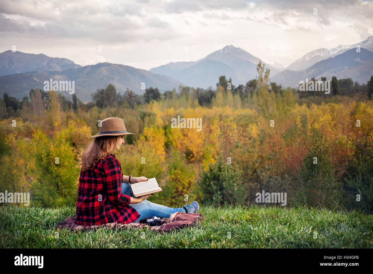 Frau in rot Hemd und Hut sitzt auf dem Rasen Lesung Buch im Herbst Wald und Berge Hintergrund überprüft Stockfoto