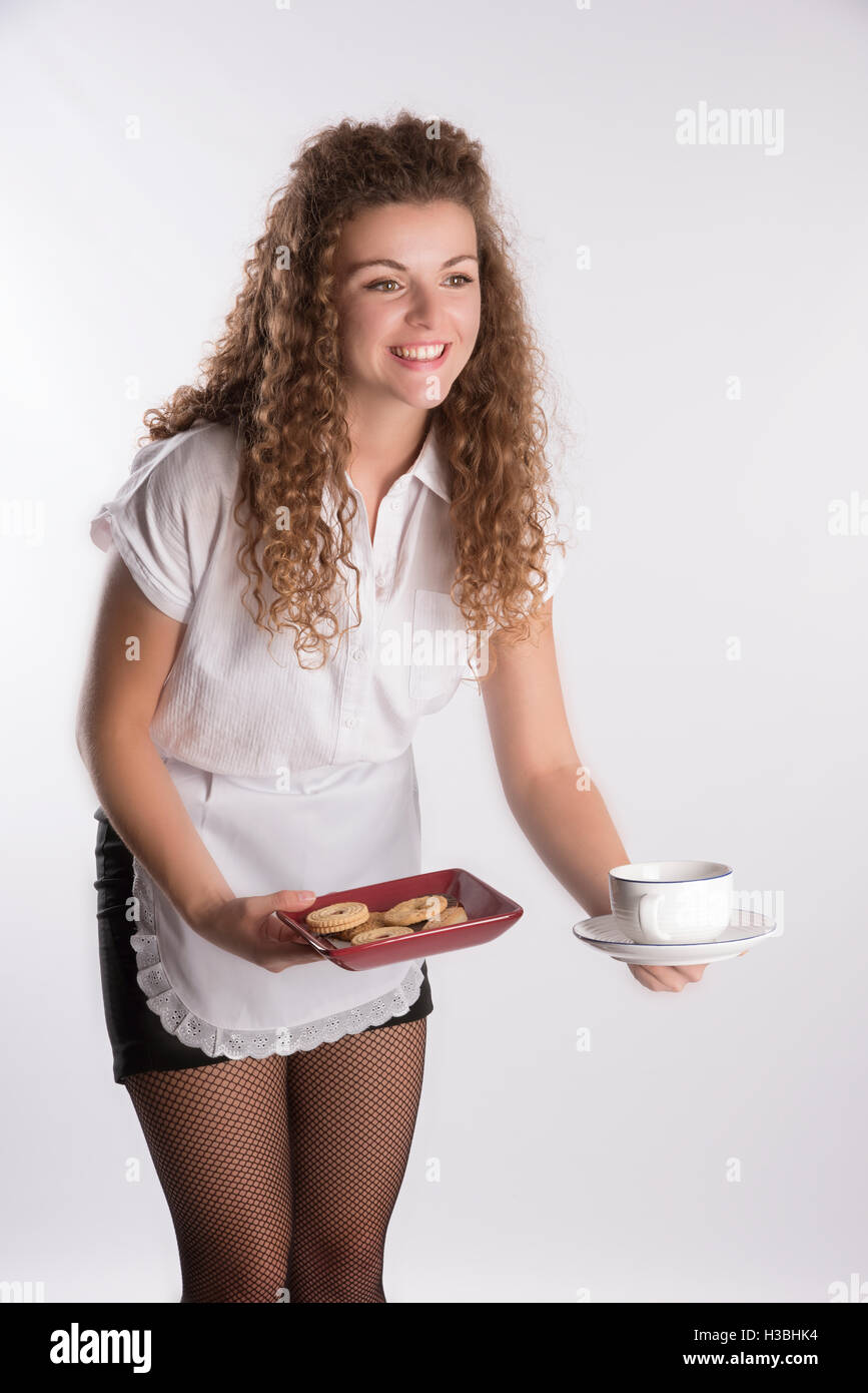 Junge attraktive Kellnerin in Uniform.  Einen langen lockigen Haaren Waitron eine Tasse Tee mit einer Auswahl an Gebäck auf einem Tablett serviert Stockfoto