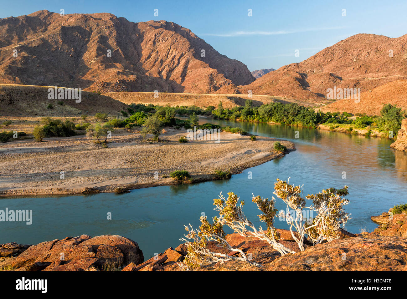 Ein Blick von Namibia nach Angola, über den Kunene-Fluss.  Atemberaubende Bett Blick auf den Fluss, mit einsamen Bergen im Hintergrund und Vordergrund Pflanzen. Stockfoto