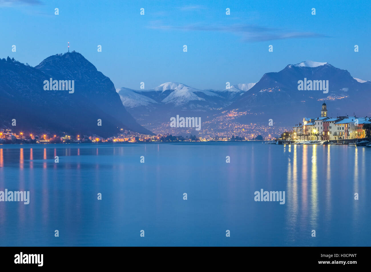 Blaue Stunde in Brusino Arsizio und Lugano in der Ferne, See Ceresio, Kanton Tessin, Schweiz. Stockfoto