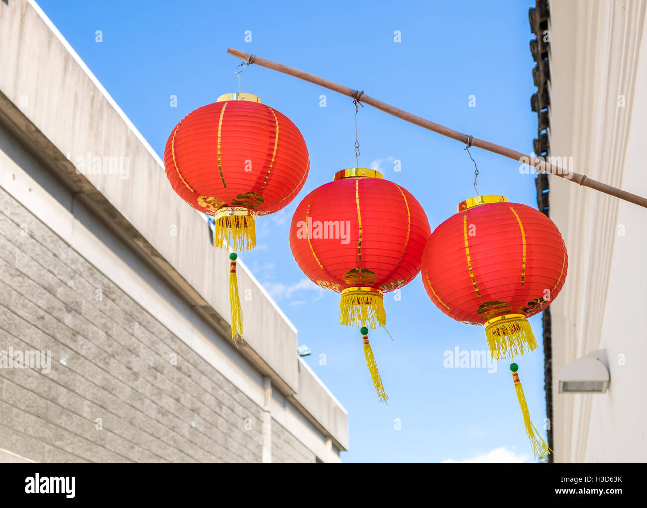 Drei rote Lampions mit goldenen Quasten hängen von einer Bambusstange außerhalb eines Gebäudes an einem Tag blauer Himmel. Stockfoto