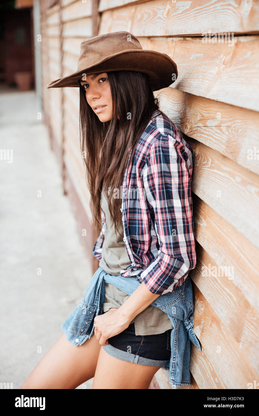 Schöne junge Frau Cowgirl in Hut und Plaid Shirt stehen in der Nähe von Haus aus Holz Stockfoto
