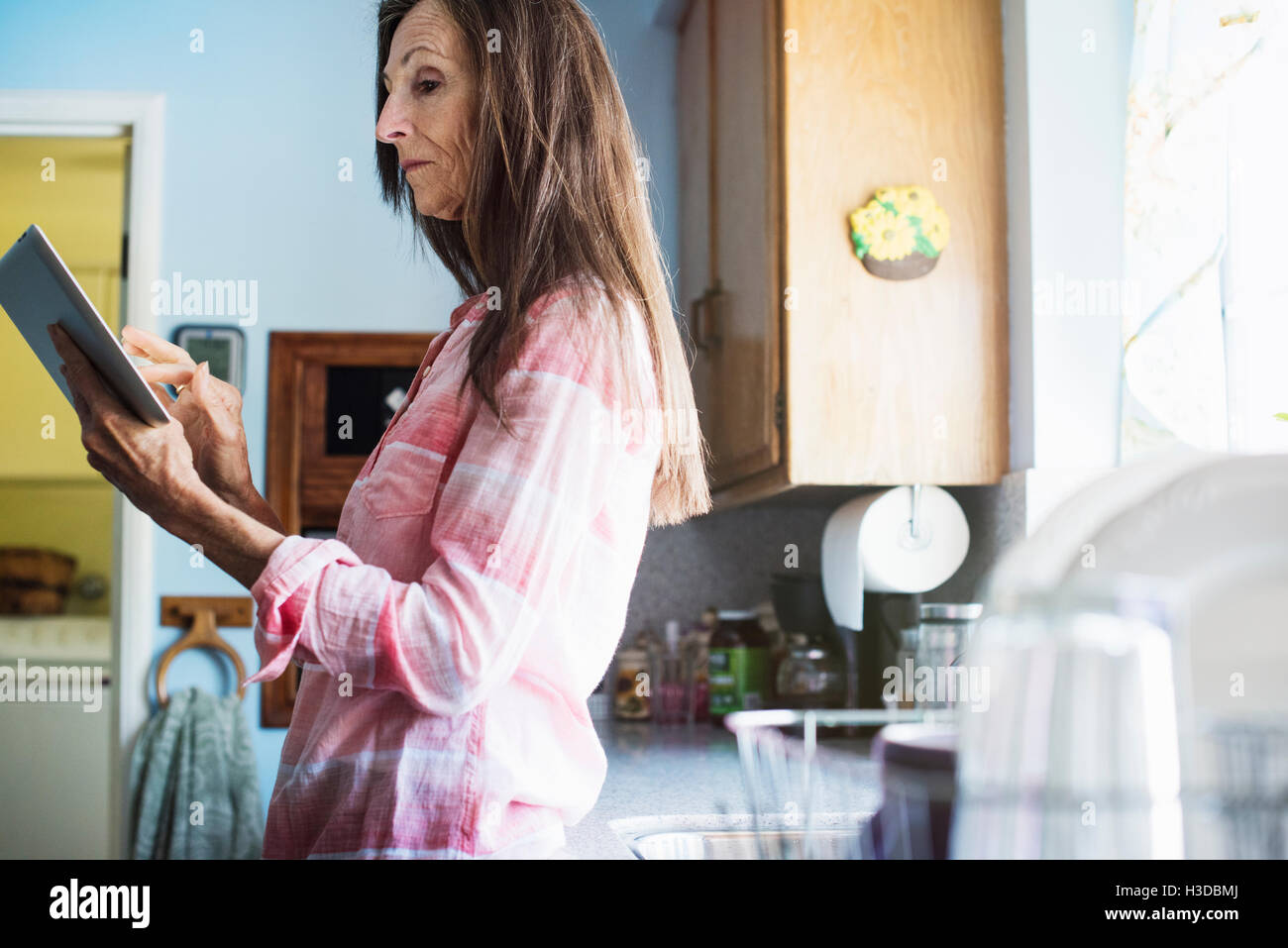 Ältere Frau mit langen braunen Haaren in der Küche stehen, mit Hilfe einer digitalen-Tablets. Stockfoto