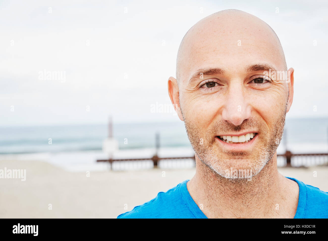 Glatze reifer Mann stehend an einem Strand in die Kamera Lächeln. Stockfoto