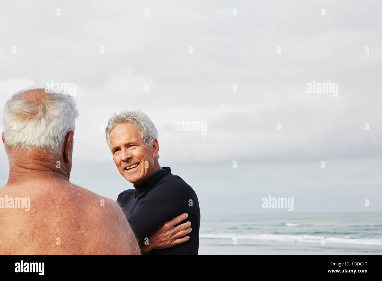 Zwei ältere Männer stehen an einem Strand im Chat, einen Neoprenanzug tragen. Stockfoto