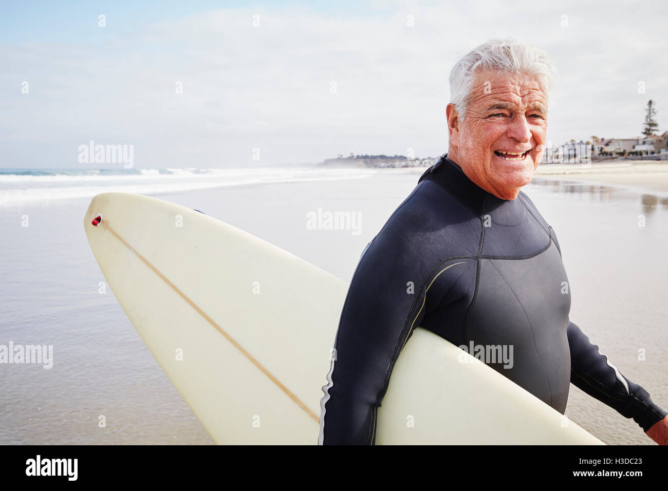 Lächelnd senior Mann stehend an einem Strand, einen Neoprenanzug tragen und tragen ein Surfbrett. Stockfoto