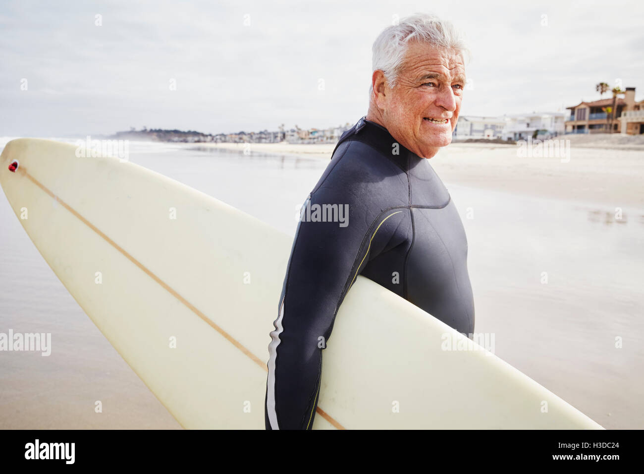 Lächelnd senior Mann stehend an einem Strand, einen Neoprenanzug tragen und tragen ein Surfbrett. Stockfoto