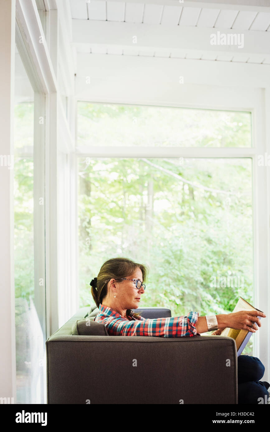Eine Frau in ein kariertes Hemd mit Brille sitzt auf dem Sofa ein Buch zu lesen. Stockfoto