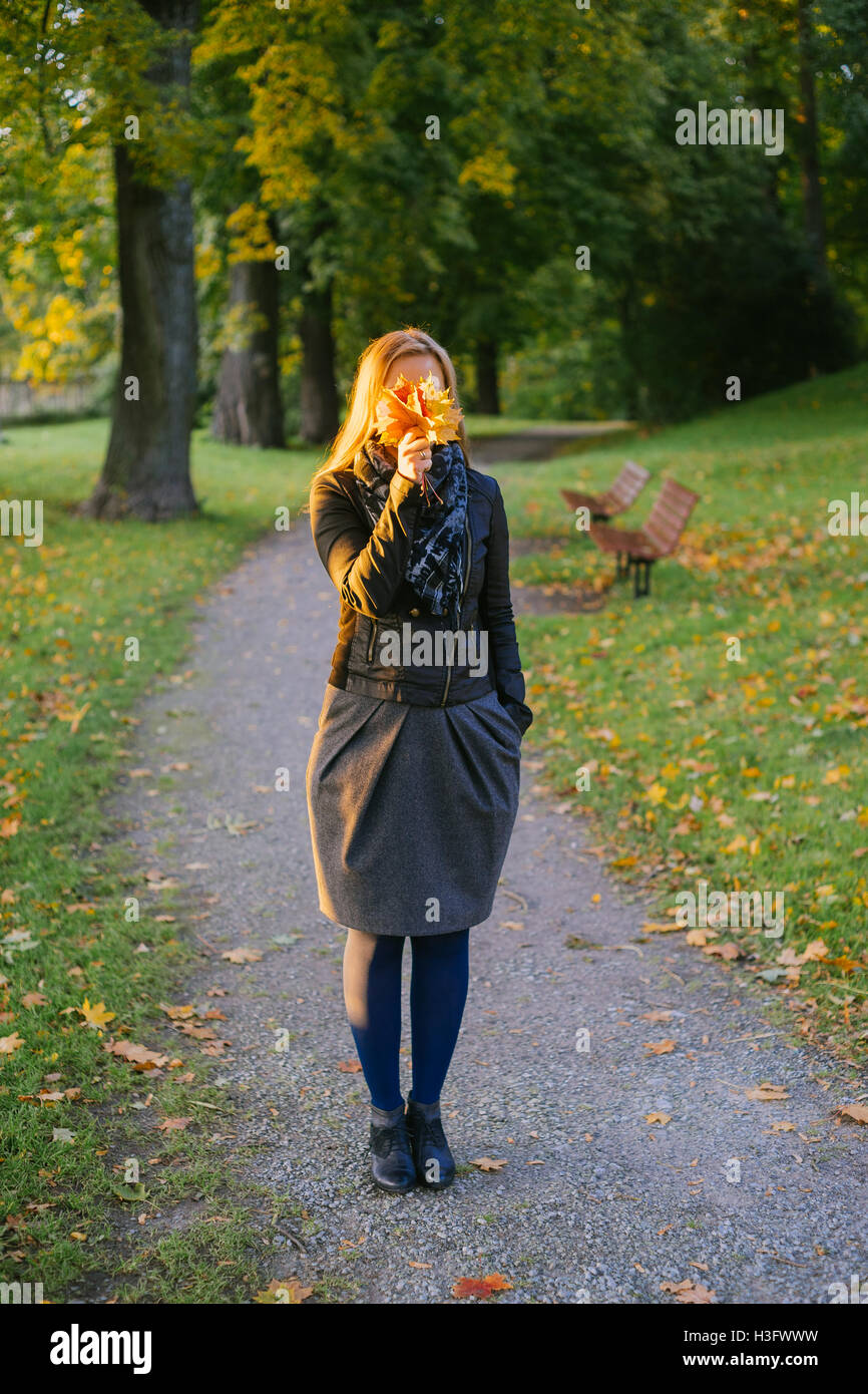 Junge Dame stehend auf Pfad im herbstlichen Park und Gesicht hinter Haufen Laub versteckt Stockfoto