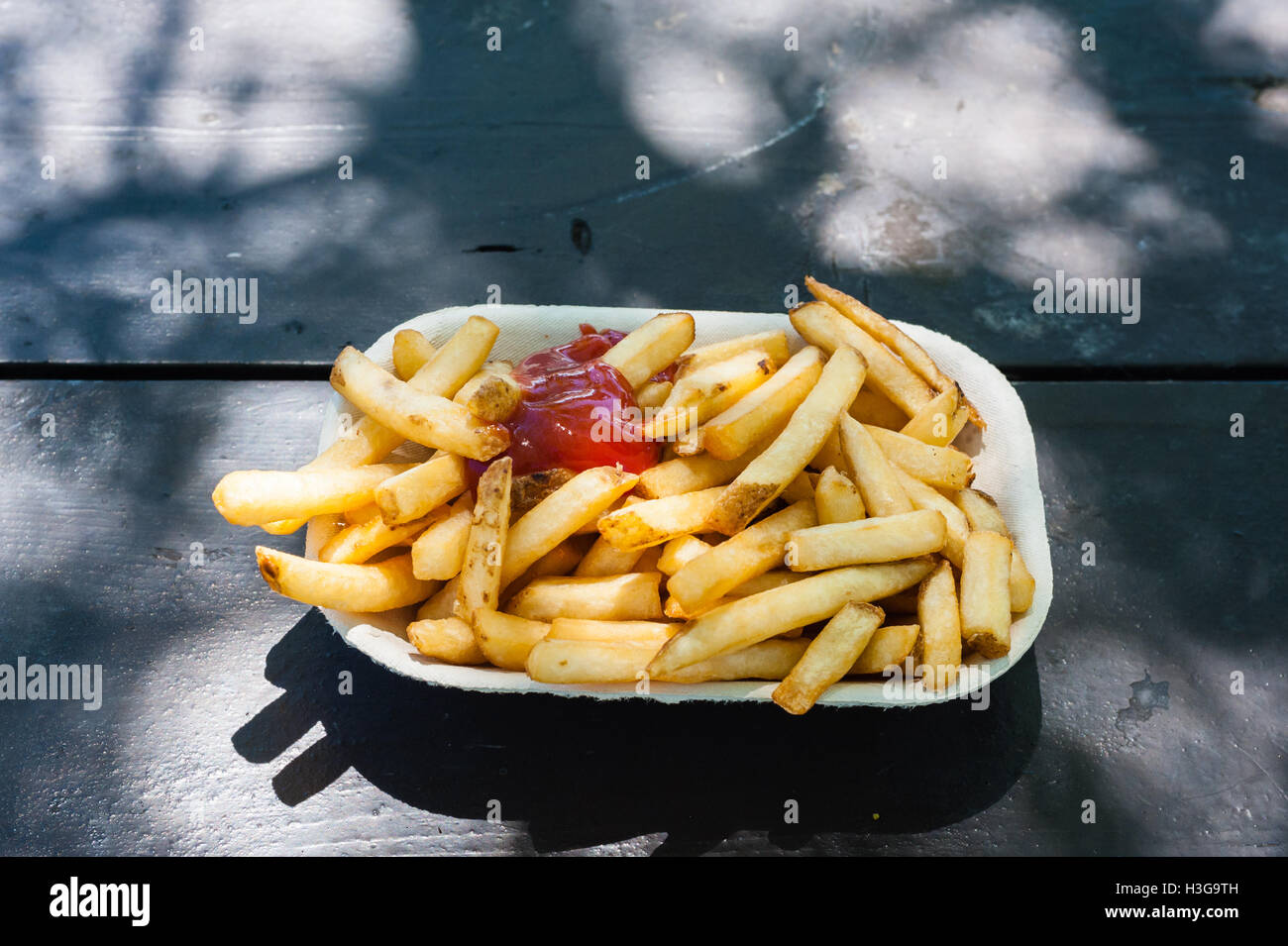 Frische Pommes Frites mit Ketchup in Papier-Container auf Holztisch in teilweise Licht und Schatten im Freien, von oben gesehen. Stockfoto