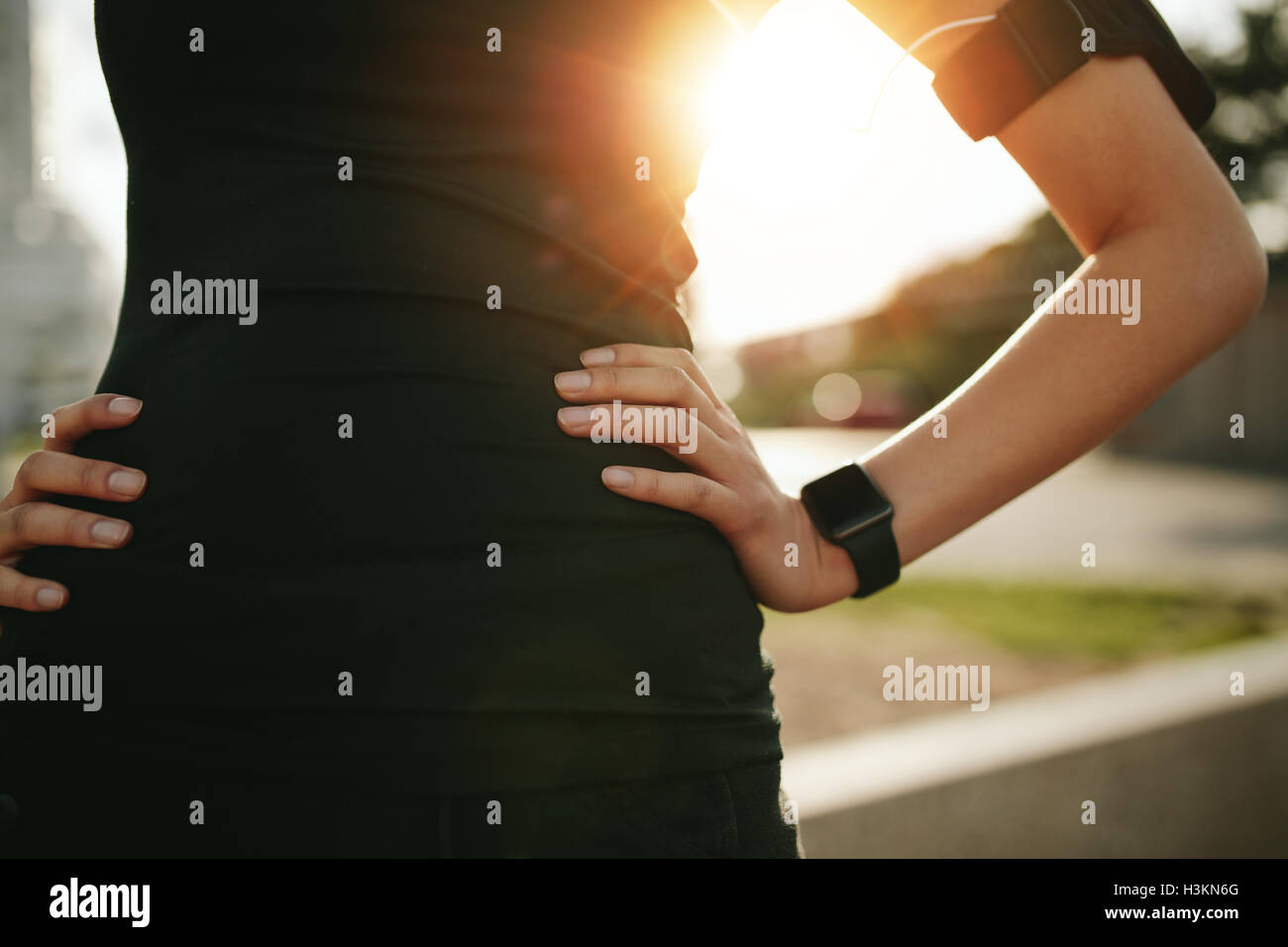 Zugeschnittene Aufnahme Fit Frau im Sport tragen stehend mit der Hand auf der Hüfte im Freien, mit Sonne Flare. Sportlerin Smartwatch tragen Stockfoto