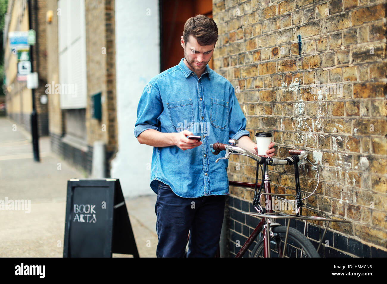 Junger Mann hält Kaffee und Handy mit Push Bike Check lehnte sich gegen die Mauer Stockfoto