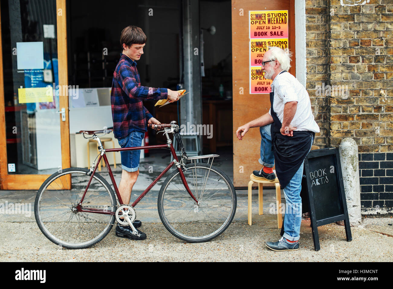 Junger Mann mit Laufrad mit Blick auf Post und stehend mit senior Handwerker außerhalb Druckwerkstatt Stockfoto