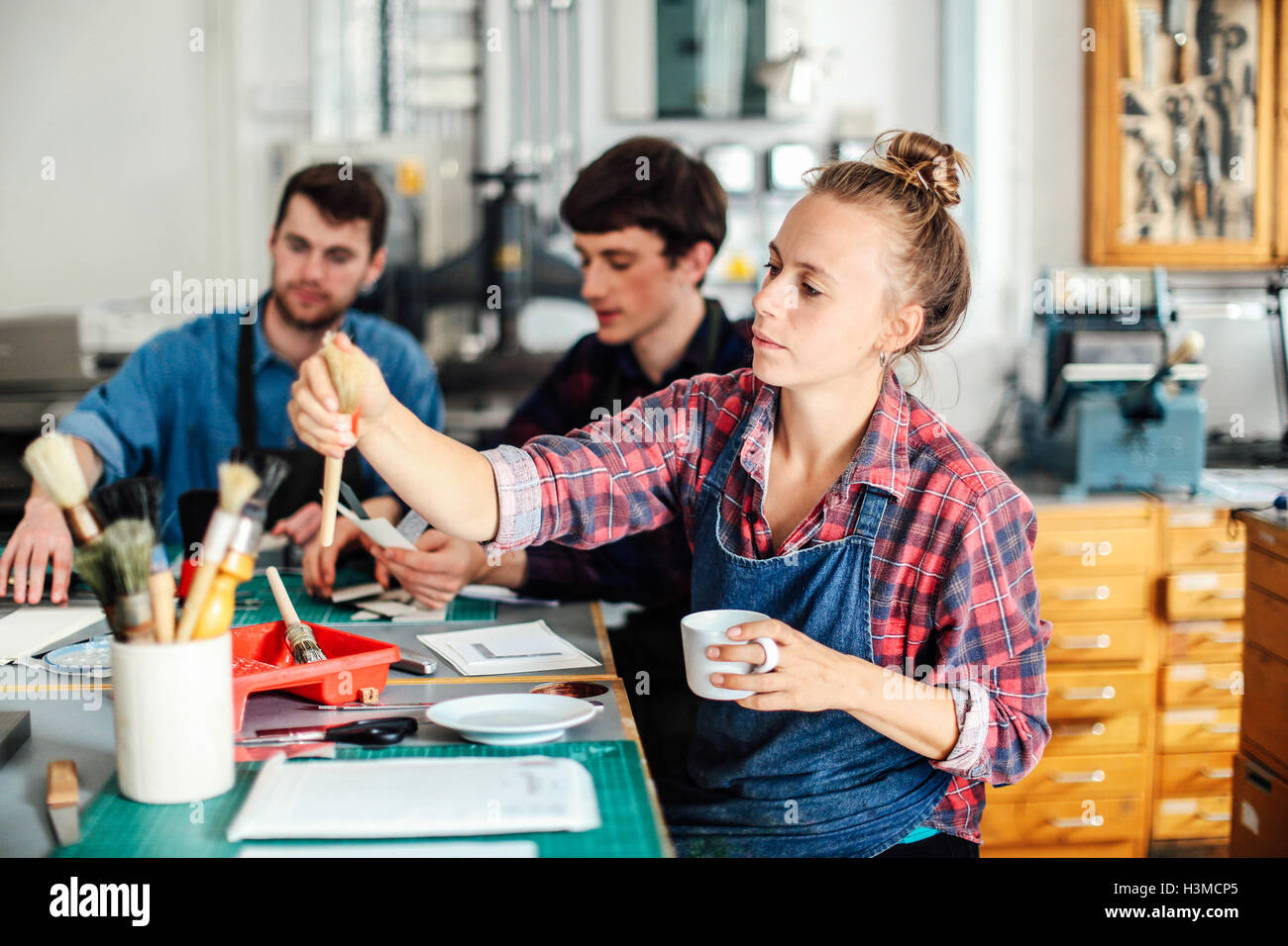 Junge Handwerkerin Kaffeetasse halten und Griff nach Pinsel in kreative print Studio, mit zwei Handwerkern im Hintergrund Stockfoto