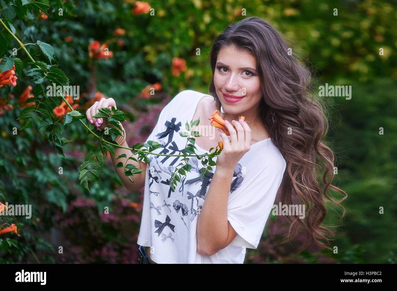 Schöne junge Frau mit langen lockigen Haaren posiert in der Nähe von Blumen in einem Garten Stockfoto