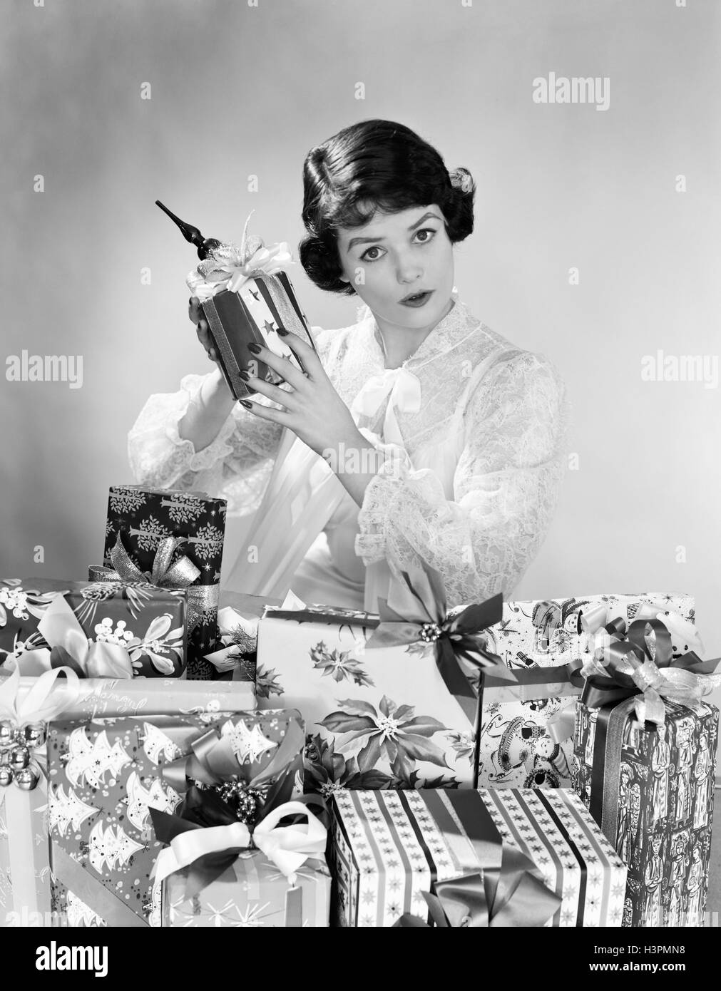 1950ER JAHREN FRAU TRAGEN NACHTHEMD WEIHNACHTSGESCHENKE MORGEN SITZEN VON HAUFEN VON WEIHNACHTEN HOLDING SCHÜTTELN EIN GESCHENK Stockfoto