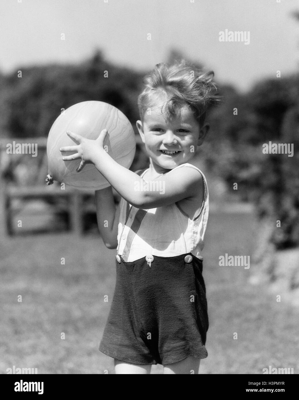 1930ER JAHREN LÄCHELNDE JUNGE IM SOMMER IM FREIEN SPIELEN IM GARTEN, BLICK IN DIE KAMERA BEREIT, TOSS BALL Stockfoto