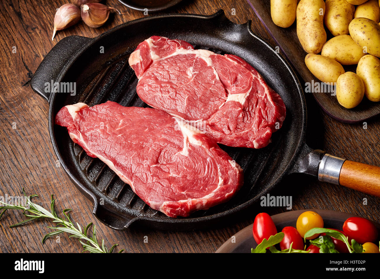 Rohes Rindfleischsteak auf dem Grillpfanne, Kartoffeln, Gewürze und Tomaten auf Holztisch Stockfoto