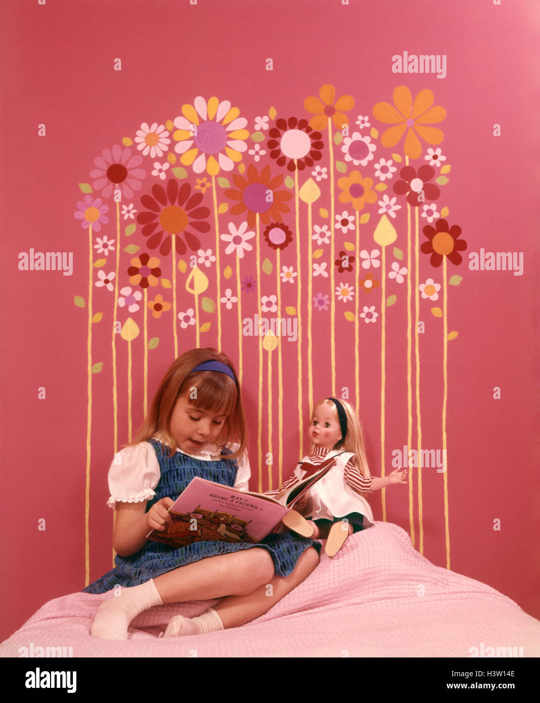 1970ER JAHREN GEMACHT MÄDCHEN SITZEN BETT LESEN ZU DOLL ROSA WÄNDE SCHLAFZIMMER KOPFTEIL DER BUNTEN BLÜTENFORMEN AN WAND GEKLEBT Stockfoto
