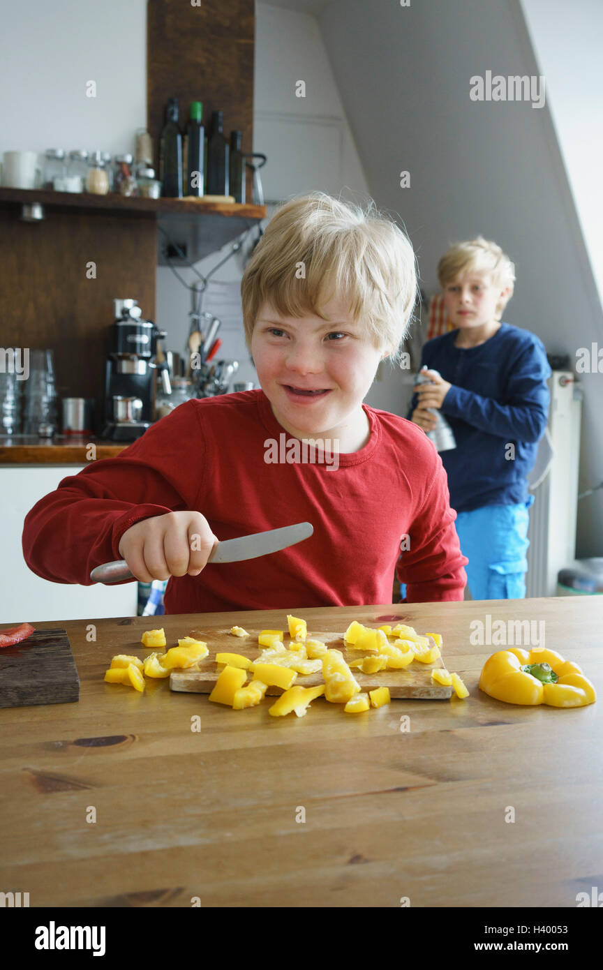 Glücklich behinderten jungen hacken gelbe Paprika mit Bruder im Hintergrund in Küche Stockfoto