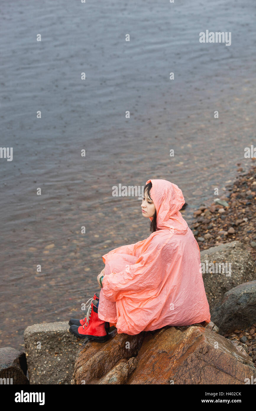 Nachdenkliche Frau mit Regenmantel sitzt auf Felsen am Ufer in der Regenzeit Stockfoto