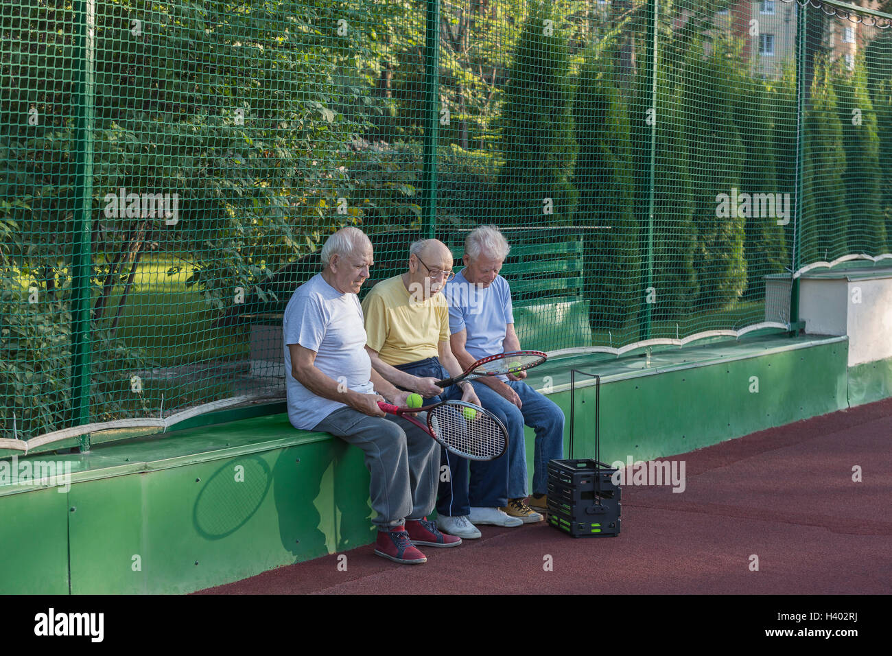 Senioren Freunde mit Tennisschläger sitzen gegen Zaun am Hof Stockfoto