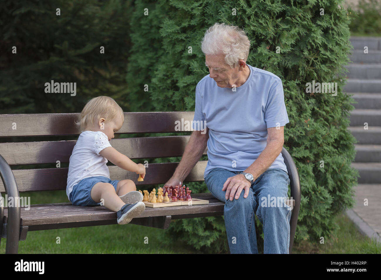 Ältere Mann spielt Schach mit Enkelin auf Parkbank gegen Baum Stockfoto